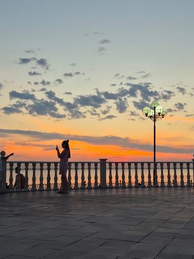 The image depicts a sunset scene along a paved walkway, possibly a promenade or boardwalk. The sky is ablaze with vibrant orange and yellow hues, indicative of the golden hour. Scattered clouds add texture to the sky.
A woman, silhouetted against the sunset, stands on the right side of the image, looking down at her phone. She is wearing a striped dress. To her left, a decorative lamppost with multiple globe-shaped lights stands prominently. A dark, ornate railing runs along the edge of the walkway, separating it from what appears to be a body of water or a lower level beyond. The ground is paved with large, dark tiles. In the lower portion of the image, a partial silhouette of another person can be seen. The overall mood is serene and peaceful, capturing the beauty of a sunset. The location is Kokshetau, Kazakhstan.