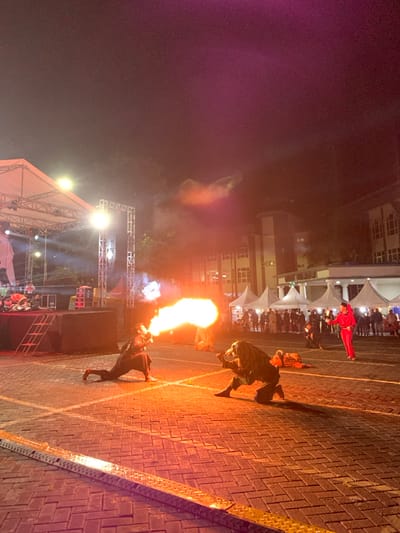 The image shows a performance of fire breathing in the evening at an outdoor event in the city of Batu, Indonesia. The location is identified by the coordinates -7.867 latitude and 112.514 longitude. The performance takes place on a brick-paved square in front of a stage, surrounded by white tents. There are two fire breathers performing. The performer on the left is breathing fire, and the performer on the right is kneeling in a dramatic pose, with a large flame visible in the background. The performer on the left wears a black costume and a black hat. The performer on the right wears a dark green costume. In the background, there are people watching the performance, and several white tents, illuminated by white lights. There is a large stage to the left, partially visible, with a large white awning stretched over it. The sky is dark, and there is smoke in the air.