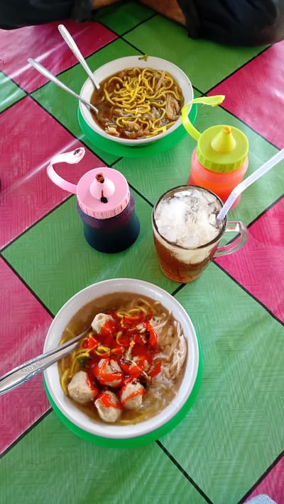 An overhead shot shows two bowls of noodles, a glass of iced tea, and two squeeze bottles on a green and pink checkered tablecloth. The bowl in the foreground contains noodles, meatballs, and a red sauce. The bowl in the background also contains noodles and broth. The glass of iced tea is filled with ice and a straw. One squeeze bottle is pink with a black lid, and the other is orange with a green lid and a green spout. The scene appears to be set in an outdoor dining area in Cotkumbang, Indonesia, possibly at a street food stall or a casual eatery. The lighting suggests it is daytime. No people are visible in the image.