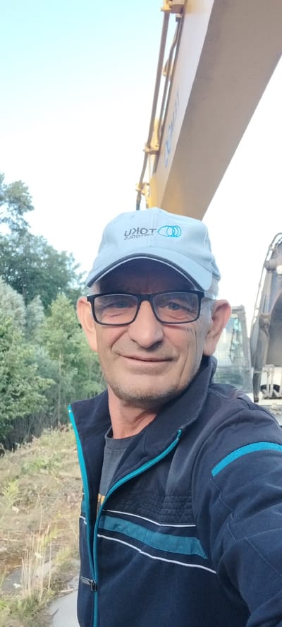 A man in a light blue baseball cap and glasses is taking a selfie outdoors. The man is wearing a dark blue jacket with turquoise and white stripes. The background shows trees and a yellow excavator. The image appears to have been taken in Novoselytsia, Ukraine.