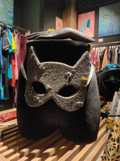 In Baan Mae Nam, Thailand, a black mannequin leg wears a textured gray cat mask. The mask has two eye holes and pointed ears. A gold chain is draped around the mannequin's neck and over its shoulder. Behind the mannequin, racks of colorful clothing are visible. The mannequin stands on a wooden surface.
