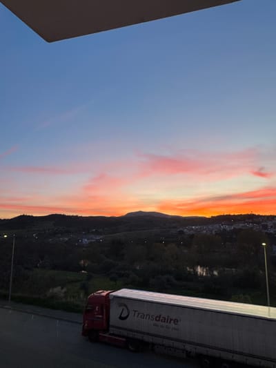 The image depicts a truck with the logo "Transdaire - We do for you" parked on a road in Mirandela, Portugal. Behind the truck, there's a cityscape bordering what appears to be a forested or mountainous area, illuminated by streetlights. The sky is a gradient of blue and orange, suggesting either sunrise or sunset. The perspective suggests the photo was taken from an elevated position, potentially from a building or higher ground nearby. The scene is captured at dusk or dawn given the lighting conditions. A corner of a building appears to be visible in the left corner of the image.