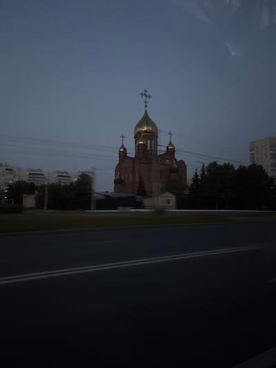 This image captures a brick church with golden domes at dusk in the city of Kemerovo, Russia. The church, likely an Orthodox Christian place of worship, is centrally located in the frame, illuminated by faint ambient light. Its distinctive onion domes and crosses are visible against the darkening sky. To the left and right of the church, apartment buildings and trees line the horizon, suggesting an urban setting. A road with white lane markings runs across the foreground, indicating the photograph was taken from a moving vehicle. The overall atmosphere is calm and serene, characteristic of twilight in a city.
