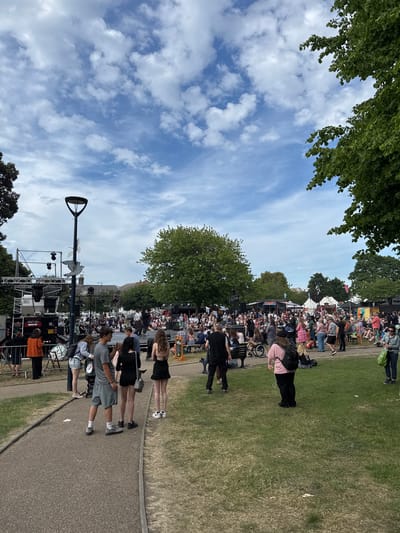 This photograph captures a daytime outdoor event in Great Yarmouth, United Kingdom. A crowd of people, appearing diverse in age and attire, are gathered on a grassy area. Some are seated on chairs or on the ground, while others are standing and walking. A stage is visible in the lower portion of the image, along with sound equipment and scaffolding, suggesting a performance or event. A prominent lamppost stands near the stage, featuring a bird sculpture at its top. The sky is partially cloudy with areas of blue. Trees are visible at the periphery of the frame. A paved path runs along the bottom of the frame, with several people standing and walking on it.