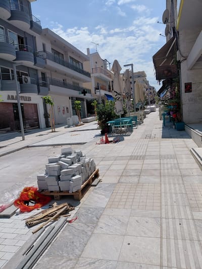 This is a street scene in Aigio, Greece, on a bright, sunny day with scattered clouds. The image captures a street undergoing renovation or construction. In the foreground, a stack of light-colored, rectangular paving stones rests on a wooden pallet. Adjacent to the pallet are scattered pieces of wood, some plastic sheeting, and a bright orange mesh bag.
The street is paved with large, light-colored marble tiles, some of which have a textured, tactile paving pattern for the visually impaired. The sidewalks are also paved with similar tiles. On the left side of the street, a building with multiple balconies, painted in gray and white, is visible. The ground floor of this building appears to be a shop with a closed metal shutter and a sign that reads "AMARYLLIS."
Further down the street, on the right side, are outdoor seating areas with tables and chairs, suggesting a cafe or restaurant. Some tables are covered with umbrellas. The buildings lining the street are multi-story structures, typical of urban environments. There are no people visible in the immediate foreground or midground. The overall impression is one of ongoing work and preparation for reopening or refurbishment.