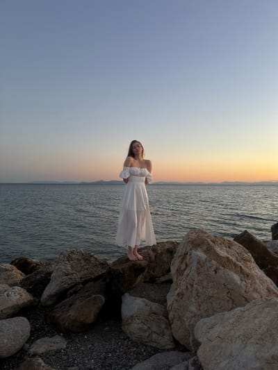 A young woman stands on a rocky shore in Paleo Faliro, Greece, wearing a white off-the-shoulder dress. The sea is calm behind her, with a distant coastline visible on the horizon. The sky displays a gradient of colors, transitioning from a pale blue at the top to a warm orange and yellow near the horizon, indicating either sunrise or sunset. She is positioned centrally in the frame, looking towards the camera with her hands clasped behind her back. The rocks in the foreground are large and varied in color and texture, with pebbles and smaller stones filling the spaces between them. The overall scene evokes a serene and picturesque atmosphere.
