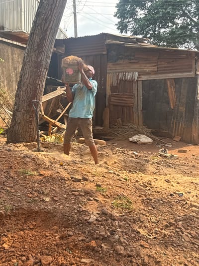 An adult male is observed performing manual labor outdoors in Aguata, Nigeria. He stands in a reddish-brown earthen depression, centrally positioned, and is actively lifting a white plastic bucket full of reddish-brown soil above his head. He wears a light blue short-sleeved shirt, brown shorts, and a light-colored cap, with a light blue facial covering visible below his nose. His skin and clothing are dusted with reddish-brown earth. To his left, a large tree trunk is visible, with a dark-handled shovel leaning against its base. In the background, behind and to the right of the man, stands a structure constructed from corrugated metal sheets and wooden planks, exhibiting rust and weathering. Scattered on the ground near this structure are palm fronds, a white bag, and miscellaneous debris. The foreground consists of uneven, rocky reddish-brown soil with minimal green vegetation. The sky is partially overcast.