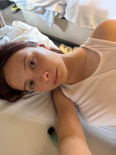 A young woman with auburn hair lies on her side on a bed with white sheets. She is wearing a white ribbed tank top. Her head is tilted, and she is looking towards the camera with green eyes and a slight smile. Next to her on the bed is a small, teal-colored vape device. In the background, a pair of yellow clogs and a pair of athletic shoes are visible on the floor. The setting appears to be a bedroom.
