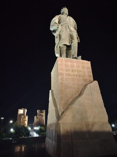 A low-angle, full-shot image taken at night shows a large bronze statue of Amir Temur, a historical conqueror, standing atop a stone pedestal. The pedestal is inscribed with the words "AMIR TEMUR" in raised letters. In the background, two illuminated ancient structures, resembling minarets or towers, stand against a dark sky. Streetlights and trees are visible in the middle ground, and reflections can be seen on a wet surface in the foreground, likely a plaza or courtyard. The overall scene is illuminated by artificial lights, creating dramatic shadows.