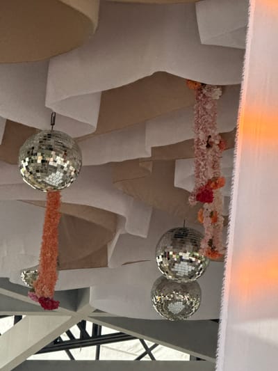 The scene depicts an indoor setting with decorative elements suspended from the ceiling. There are at least four disco balls of varying sizes and two garlands of flowers, one in shades of pink, peach, and red, and the other in peach and red. The backdrop consists of a series of draped, layered fabrics in white and tan. In the upper portion of the frame, there is a horizontal expanse of white fabric, suggesting a ceiling or overhead structure. Visible in the right corner are structural support beams, possibly part of the building's framework.

Given the style of decoration and architecture, and based on visual cues, it is possible this scene is located in a public or event space in Santiago de Querétaro, Mexico. However, without more precise information, it is impossible to verify the actual place.
