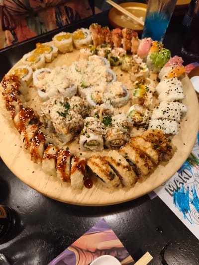 The image shows a large wooden platter of sushi, predominantly rolls, arranged circularly on a dark, presumably black, table. The sushi varieties include those topped with mango, fried rolls drizzled with sauce and sprinkled with sesame seeds, rolls with a shredded topping, rolls with avocado and other toppings, and rolls sprinkled with black sesame seeds. Behind the platter, a blue drink in a tall glass is visible, along with a small bowl, chopsticks, and other items out of focus. In the lower part of the frame, there's a soy sauce bottle, chopsticks, and a piece of paper with the word "MAKI" and some drawings. The scene appears to be indoors, possibly a restaurant. There is no explicit indication of being in Quito, Ecuador, in the provided image. Details about people are minimal; only part of a person's outfit is visible.