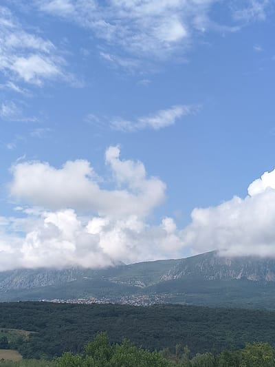 This media file depicts a natural landscape scene with a clear blue sky dotted with fluffy white clouds. The right side of the image is dominated by a densely forested mountainside, with a small village nestled along its lower slopes. The trees are a vibrant green, indicating a time of year with lush vegetation, likely spring or summer. The village appears to be situated in a valley, with buildings clustered together. The mountains are steep and rugged, with some rocky outcrops visible through the trees and clouds. The overall impression is one of a serene and picturesque rural setting. The weather appears to be pleasant and sunny, with the clouds suggesting a light breeze. The time of day is likely midday or early afternoon, given the bright sunlight. There is no visible text in the image. The scene evokes a sense of tranquility and natural beauty, characteristic of mountainous regions.