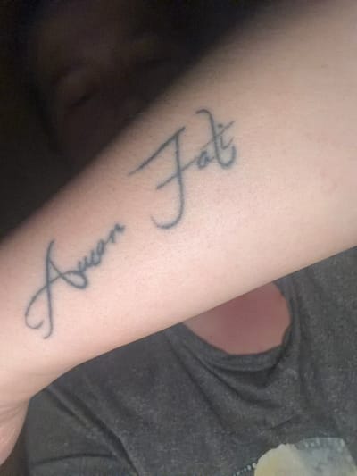 The image shows a close-up of a person's forearm with a tattoo. The tattoo consists of cursive writing. The first word appears to be "Amor" and the second word appears to be "Fati". The forearm is positioned against a dark grey t-shirt. The background is dark and out of focus, obscuring any environmental details. There is no information in the image to suggest a location in Szczecin, Poland.