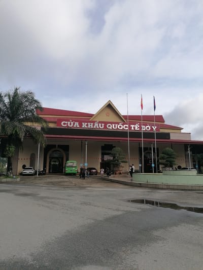 The image depicts the Bo Y International Border Gate in Bo Y, Vietnam. The border gate building is a large, modern structure with a red-tiled roof and beige walls. Large white lettering on the building reads "CỬA KHẨU QUỐC TẾ BỜ Y". In front of the building, there is a paved area with several vehicles, including a white sedan, a green minibus, and a red sedan. A few people are visible, some walking, and some standing near the vehicles. Two flags, one red with a yellow star (Vietnam) and one blue with a white symbol (likely Laos), are flying from poles in front of the building. To the left, a tall palm tree stands beside the building. The sky is cloudy. The ground in the foreground is wet, indicating recent rain, with puddles reflecting the scene.