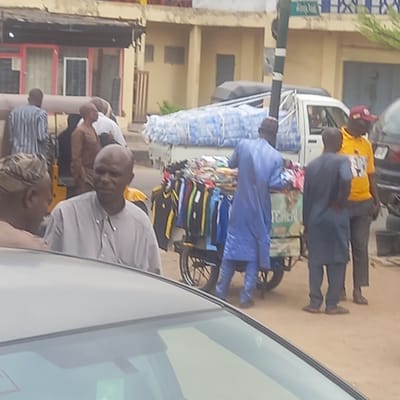 The scene depicts a street in Dadi Riba, Nigeria. Several individuals are visible, including men dressed in casual attire and traditional blue robes. One man is pushing a cart loaded with bottled water and colorful clothing. The background features a building with windows and a nearby sign. The image appears to be taken from inside a vehicle, with part of the car's interior visible. The environment suggests a typical urban scene with commercial activity.