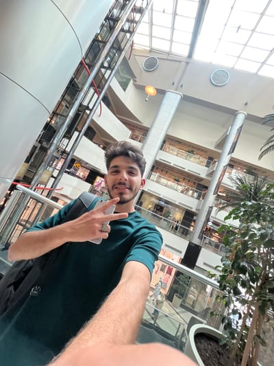 A young man is captured in a selfie within a large, brightly lit indoor complex, likely a shopping mall in Kafr Sousa Municipality, Syria. He smiles directly at the camera, holding a light-colored smartphone in his left hand while making a 'V' or peace sign with his right hand. He wears a dark teal short-sleeved t-shirt and a distinct, intricate ring on his left ring finger. A black backpack is visible over his left shoulder.
The background reveals a multi-story modern building with several levels of commercial spaces featuring glass facades. Prominent architectural elements include shiny metal escalators or glass elevators with red railing accents on the left side. The structure benefits from ample natural light streaming through large overhead windows or skylights. Overhead, large cylindrical structures, possibly for advertisements, are suspended, and round metallic grilles are visible on the ceiling. The space is adorned with large potted plants, including palm trees and leafy green foliage, contributing to a vibrant indoor atmosphere. Faint text, possibly "ELLE," can be discerned on the signage of one of the shops in the background. The scene depicts a bustling commercial environment during the daytime.