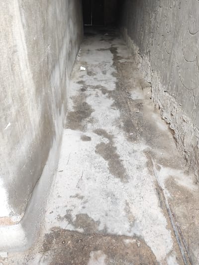 The image displays a narrow, concrete alleyway or corridor located within the city of Al-Qanawat Municipality, Syria. The scene is viewed from an elevated perspective, looking down the length of the passage into an unlit or dimly lit area, where a dark rectangular opening or door is barely discernible at the far end. The left wall is constructed from smooth, light gray concrete, exhibiting extensive dark, damp stains and streaks, particularly towards its base. The right wall features a dark gray, textured concrete surface with a repetitive, raised, wave-like or scale-like pattern. Its lower section is rough and appears partially eroded or unfinished. The concrete floor is a light gray, heavily marked with irregular dark, wet patches and accumulated dirt. A thin, dark linear object, resembling a metal pipe or wire, runs along the base of the right wall on the floor. A single white, small paper or plastic cup lies discarded on the floor towards the left side of the passage. There are no individuals, visible actions, or interactions present in the scene.