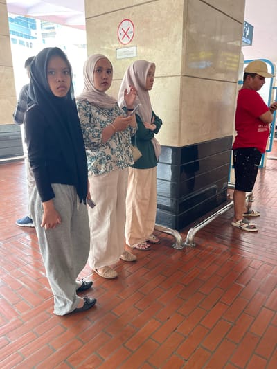In Tangerang, Indonesia, a group of four people are standing in line on a reddish-brown tiled floor. The first person in line, from the left, is a young girl wearing a black hijab and grey sweatpants. The second person is a woman in a light-colored hijab and a floral patterned blouse, holding a smartphone. The third person is a woman wearing a light purple hijab and a green jacket, also with a handbag. The fourth person, on the far right, is a man wearing a red t-shirt, black patterned shorts, and a yellow baseball cap. He is looking at his phone. Behind the people, there is a large pillar with beige marble-like cladding. A silver railing runs along the floor in front of the pillar. Above the pillar, a sign with an information symbol and text is visible. Another sign with a red circle and a diagonal line through a cigarette symbol indicates a no-smoking area. The environment appears to be an indoor public space, possibly a transportation hub or a shopping mall.