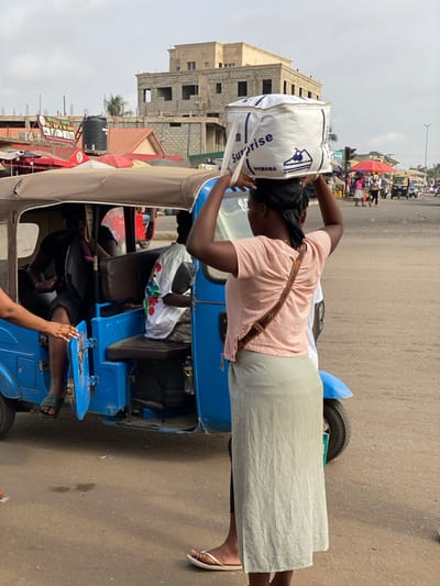 The scene depicts a daylight street view in Ablekuma, Ghana. A person, observed from the rear, stands or walks in the foreground, balancing a large white rectangular bag labeled "Surprise" with a cake graphic on their head. The individual wears a pink short-sleeved top with a gathered back detail, a light-colored patterned long skirt, and white flip-flops, carrying a brown shoulder bag featuring an 'LV' monogram. Adjacent to this individual, a blue three-wheeled motor vehicle, equipped with a brown canopy roof, has its left rear passenger door open. Two passengers are visible inside: one in a dark shirt and shorts, with their right hand extended towards the open door, and another in a white short-sleeved shirt with a colorful pattern. The ground is a paved road surface. In the mid-ground, a multi-story concrete building is under construction, displaying exposed blockwork and rebar. Below this structure, red and beige market awnings or umbrellas are present, along with an elevated black water tank and a yellow sign for 'TWO TOPS CO LTD'. The background includes additional indistinct figures, vehicles, and urban infrastructure under an overcast sky.