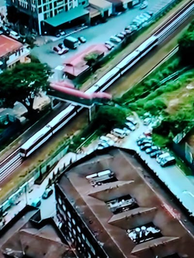 An elevated, slightly blurry, wide-angle view looks down upon a cityscape with a train moving on tracks. In the foreground, a brown, multi-leveled structure with visible openings is prominent. Further back, a light-colored train with dark windows travels along parallel tracks. Adjacent to the tracks, a strip of green vegetation is visible, and beyond that, a road with numerous parked cars. In the upper left quadrant of the image, buildings of various heights and colors, including one with a teal roof, are situated. The overall scene depicts an urban environment with transportation infrastructure and surrounding buildings. The provided location, Cherepovets, Russia, is not verifiable from the visual information present in the image.