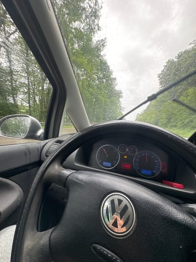The image captures a driver's perspective from inside a car in Steinalben, Germany, during overcast and rainy conditions. Raindrops are visible on the wet windshield and side mirror, through which trees and a cloudy sky can be seen. The general setting appears to be a road or parked location surrounded by nature, likely during daytime given the ambient light, though no direct sunlight is present.
The interior of the car features a black steering wheel with a visible Volkswagen (VW) logo at its center and the word "AIRBAG" partially visible. A black, textured steering wheel cover is also in place. The dashboard is visible behind the steering wheel, displaying car gauges with blue backlighting.
Notable details from the dashboard include:
* An external temperature reading of "13.0°C."
* An odometer displaying "277318."
* Other numerical readings including "964" and "653" (likely trip meters or other vehicle data).
* Speedometer markings ranging up to "260."
* An orange warning light is illuminated on the dashboard, typically indicating a tire pressure issue or a general warning.
No people are visible in the image. The scene suggests the car is either driving or parked in a natural, possibly rural or suburban, setting during inclement weather.