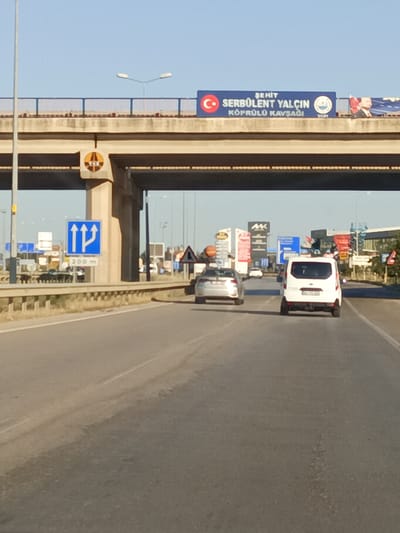 This is a daytime, eye-level, outdoor, vertical shot of a highway in Yahşihan, Turkey. The foreground shows a paved road with lane markings, suggesting a multi-lane highway. In the middle ground, a white van is driving away from the camera in the right lane. To the left of the van, a silver sedan is also driving away from the camera in the left lane. In the background, an overpass spans the highway. On the overpass, a blue banner reads "ŞEHİT SERBÜLENT YALÇIN KÖPRÜLÜ KAVŞAĞI" with Turkish flags and images of Mustafa Kemal Atatürk. Below the overpass, a blue traffic sign indicates three lanes ahead, with the left lane splitting into two. Further in the background, commercial buildings and billboards are visible. The sky is clear and blue.