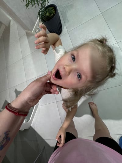 The image depicts a low-angle view of a young child with blonde hair in pigtails, looking upwards with an open mouth. A hand with a bracelet rests near the child's face. Another person is partially visible in the upper right, wearing black leggings and a pink garment. A potted plant sits on the left, and a white tiled floor covers the ground. Based on the location of Gornyi (Dvurechenskoe S/P), Russia, the setting is most likely a domestic interior.
