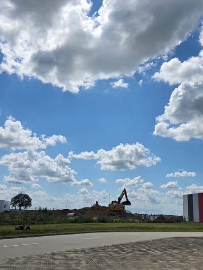 This is a vertical, medium-shot photograph captured on a sunny day with scattered clouds. The scene depicts a construction site with a yellow excavator in the foreground, positioned on a mound of dirt. The excavator appears to be in operation, with its arm extended. Behind the excavator, there is a large pile of dirt, a fence, and some green vegetation. In the distance, a building with red and white sections is visible. A paved area with white lines runs along the right side of the image, suggesting a road or parking lot. A single tree with dark green foliage stands to the left of the paved area. There is also a lawnmower visible on the grass near the tree. The sky is a vibrant blue, filled with fluffy white and gray clouds. The overall impression is one of outdoor work in progress under pleasant weather conditions. Text visible on the excavator reads "CAT".