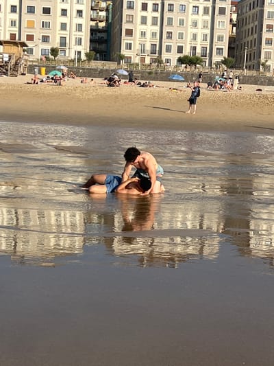 Two men are in shallow water on a beach, with one man kneeling and the other lying on his back. The kneeling man appears to be helping the lying man. The beach is sandy, with buildings in the background that suggest an urban environment. The water reflects the buildings. In the background, other people are visible on the beach, some are sitting or lying down, and others are walking. Beach umbrellas and a small lifeguard tower are also present. The scene suggests a warm day at a beach. The architecture of the buildings suggests a European city, and the presence of a beach is characteristic of coastal cities. Given the context of San Sebastián, Spain, this scene is likely La Concha beach, a well-known urban beach with a prominent promenade and buildings.