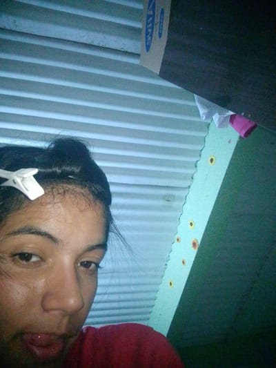A close-up, low-angle selfie shows a young woman with dark hair styled in a bun and a white hair clip on her forehead. Her eyes are dark brown, and she is looking towards the camera. Her mouth is slightly open, with her tongue visible. She is wearing a red top. The background consists of corrugated metal siding and a light blue wall adorned with several small, painted sunflowers. The scene appears to be indoors, possibly in El Maco, Venezuela, based on the provided context. There are some items of clothing, a pink and white cloth, hanging near the top of the frame.