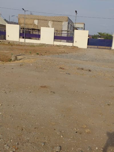 A partially constructed building with a block facade stands behind a fence of white columns and purple metal bars. A gate matches the fence's color scheme. The foreground is dominated by a dirt and gravel road, suggesting an undeveloped or rural area. Light poles are interspersed along the fence line. The sky is overcast, giving the scene a muted color palette.