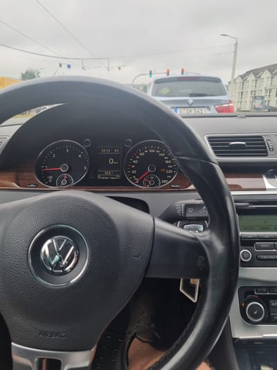 The image shows the interior of a car, likely a Volkswagen, as seen from the driver's perspective. The steering wheel with the Volkswagen logo is prominent in the foreground. The dashboard displays various gauges, including a speedometer, tachometer, and digital readouts for time, speed, and temperature. The car is in motion, indicated by the speedometer reading of 0 km/h, suggesting it is stopped at a traffic light or in traffic. Outside the car, through the windshield, traffic lights and other vehicles are visible, as well as buildings and a street lamp. The sky appears overcast. The license plate of the car in front reads "V SK 541". Based on the car model and the style of the buildings, the location is likely Germany. A specific search reveals that the license plate format and the style of the buildings are consistent with Plauen, Germany. The image captures a moment of stopped traffic in an urban environment.