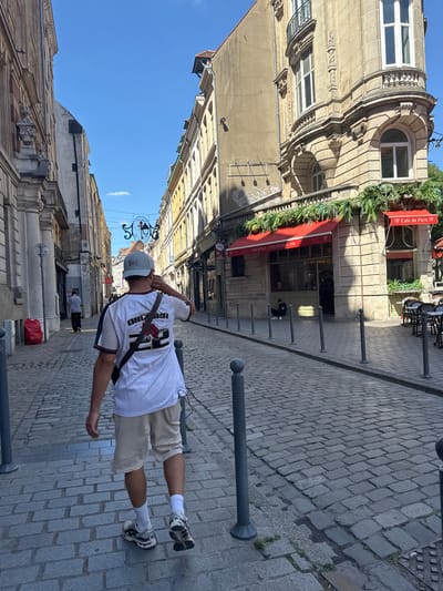 The image depicts a street scene in Lille, France. The foreground features a person, likely male, wearing a white t-shirt with a black and red design, khaki shorts, white socks, and athletic shoes. A gray baseball cap covers his head, and a dark-colored bag is slung across his chest. His right arm is extended, and he appears to be looking to his left.

The setting is a cobblestone street lined with historic buildings. To the left, a grand stone building with ornate architectural details and arched windows dominates the frame. A red awning of "Café de Paris - CHATOURY 1930" is visible, adorned with greenery, suggesting a café or restaurant. Further down the street, other buildings with varied architectural styles and some with signs are visible. Several dark bollards are placed along the sidewalk. The sky is a clear blue, indicating daytime and likely pleasant weather.

The image is taken from a low angle, with the majority of the frame oriented vertically. The person is positioned mid-street, facing away from the viewer towards the buildings. The overall impression is that of a casual stroll or a pause in an exploration of the city. No specific event is clearly taking place, but the scene suggests an ordinary day in a European city.