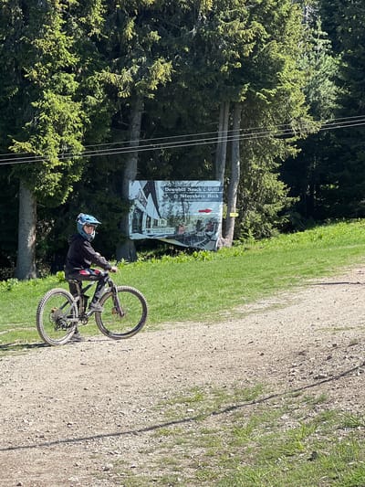A young person wearing a blue helmet and a black hoodie is sitting on a mountain bike on a gravel path. The bike has knobby tires, characteristic of off-road riding. The background consists of tall evergreen trees and a grassy slope. A large sign advertising "Downhill Snack & Grill @ Sityakovo Rock" is visible behind the person, indicating the location is likely a ski resort or a recreational area in the mountains near Samokov, Bulgaria. The ground is covered with small stones and dirt, suggesting it's a trail or path. Sunlight illuminates the scene, casting shadows.