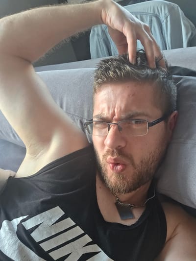 An adult male with light skin, short brown hair, and a short beard and mustache lies on a light gray upholstered surface, viewed from a high angle. He wears rectangular-framed glasses with dark rims and a silver bridge. His right arm is bent with his hand on top of his head, fingers resting in his hair. His lips are pursed in a "fish face" expression, and his eyebrows are slightly raised, creating minor furrows on his forehead. He wears a black sleeveless tank top with a faded white "NIKE" logo on the chest, and a silver-colored chain necklace with a flat, rectangular, dark metallic pendant around his neck. In the upper background, a light blue denim shirt is draped over an unseen object. The scene takes place indoors in Yagodovo, Bulgaria.