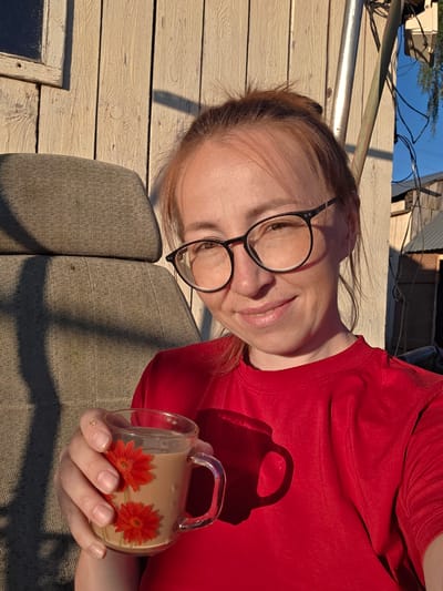 This is a close-up, rotated image of a woman lying down, holding a mug. She has light skin, reddish-brown hair, and is wearing black-rimmed glasses and a red t-shirt. Her eyes are looking towards the camera, and she has a slight smile. The mug is clear glass with a handle and contains a light brown liquid. Two red flowers are depicted on the exterior of the mug.

The setting appears to be outdoors, with wooden planks forming a surface beneath and behind the woman. Sunlight casts shadows, suggesting it is either morning or late afternoon. In the background, there are glimpses of what looks like a building with a corrugated roof and wires, along with a blue sky. The overall mood is relaxed and casual. The location context provided is "Буренка, Russia." There is no visible text in the image.