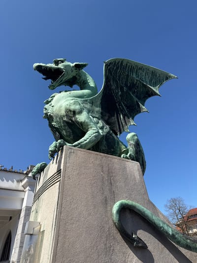 A large, imposing bronze dragon statue, exhibiting a prominent green patina, is captured from a low-angle perspective against a clear, bright blue sky in Ljubljana, Slovenia. This is one of the four dragon statues adorning the Dragon Bridge (Zmajski most) in the city center.
The dragon is depicted in a seated, vigilant posture with its powerful body facing forward and its head turned slightly to the viewer's left. Its mouth is agape, revealing interior details, and its formidable wings are spread upwards and outwards. Prominent claws grip the rough, grey stone pedestal upon which it sits. The statue's surface shows texture and vertical streaking from the patina, indicative of weathering.
The pedestal itself is a robust, rectangular structure made of light grey, possibly granite or concrete, material with carved horizontal lines and a vertical panel visible on its face. A bronze serpentine element, possibly the dragon's tail, wraps around the lower right side of the pedestal.
In the background, to the left of the pedestal, a portion of a white building with an arched window and a dark-tiled roof is visible. Several pigeons are perched along the edge of this roof. To the right, sparse, bare tree branches are visible against the sky, suggesting a non-leafy season. A rectangular plaque or sign is mounted on the left side of the pedestal, below the dragon's claws.