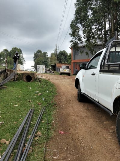 The scene depicts a semi-urban environment in Nyansiongo ward, Kenya, under an overcast sky. A winding dirt road occupies the foreground and midground, curving slightly to the right. To the immediate right, the front half of a white double-cab pickup truck, equipped with black side steps and a black roll bar, is parked or positioned on the dirt road, facing away from the camera.
On the left side of the road, a grassy verge contains several dark metal reinforcement bars lying horizontally. A red string-like object is visible on the dirt surface near the pickup truck's tire. Further along the left verge, a dark concrete culvert pipe is partially exposed in the ground. Adjacent to this, a single individual, dressed in a dark jacket, blue jeans, and a light-colored cap, stands next to a stack of rebar and wooden posts, looking downwards at an object held in their hands.
In the midground, further down the dirt road, a light-colored compact SUV or hatchback is visible, facing towards the camera. Behind the vehicles and structures, several green-leafed trees, likely eucalyptus, line the horizon. To the right of the road, a two-story building with a grey upper facade and an orange lower section is visible. Utility poles and power lines crisscross the sky. In the background on the left, white rectangular containers or utility boxes are discernible near a pile of construction debris.
