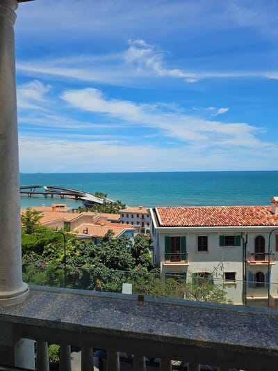 This is a vertical, wide-angle shot of a scenic coastal view, likely from a balcony. The sky, a vibrant blue with wispy white clouds, takes up the left half of the frame. The right half features a turquoise sea meeting a coastline dotted with buildings and lush greenery. In the foreground, a stone railing and a white column are visible, suggesting the vantage point.

The buildings exhibit Mediterranean-style architecture with terracotta tiled roofs and light-colored facades. Some have green shutters on their windows. Further down the coast, a modern bridge-like structure extends over the water, hinting at a resort or a pathway. Dense green trees and some flowering bushes add to the vibrant natural setting.

The bright sunlight and clear sky suggest it is daytime, likely late morning or early afternoon. The overall mood is peaceful and serene, capturing the beauty of a tropical coastal location. No text is visible in the image. The scene strongly suggests a location in Phú Quốc, Vietnam, with its characteristic coastal architecture and abundant vegetation.