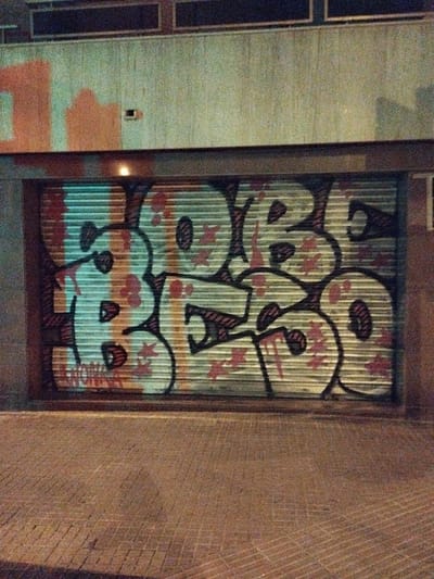 A corrugated metal garage door in Rubí, Spain, displays graffiti spelling "BOBO," outlined in dark paint and filled with light-colored paint marked with dark-red star-shaped patterns. The garage is built into a weathered facade, where remnants of older graffiti are visible in orange at the top left. The foreground shows the brick pavement of the street. The overall lighting suggests nighttime.