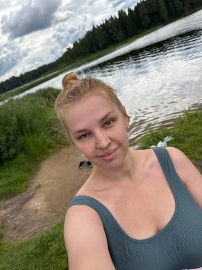 A young woman with blonde hair pulled into a bun, green eyes, and freckles is taking a selfie by a body of water. She is wearing a dark teal one-piece swimsuit. The background shows a lake with a forested shoreline under a cloudy sky. A dirt path and green grass are visible on the shore in the foreground. A pair of dark blue sandals are on the path behind her.