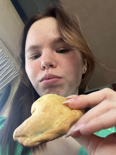 The image depicts a young female indoors in Yurkovo, Russia. Her face, viewed from a low upward angle, shows a light complexion with some facial redness and prominent brown hair with lighter highlights. A small earring is visible in her proper right ear. Her lips are slightly parted, suggesting an ongoing or imminent action of eating. She is holding a light golden-brown, irregularly shaped pastry in her proper right hand. Her fingernails are long, oval-shaped, and appear to have a natural or clear polish. She is wearing a green garment. The background consists of a blurry, light-colored ribbed panel or vent on the left and an otherwise indistinct, warm neutral-toned interior, consistent with an enclosed space.