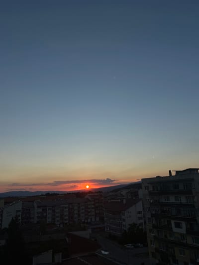 This media file depicts a sunset over a city, likely Samokov, Bulgaria. The scene is dominated by a vast expanse of sky, transitioning from a deep blue at the top to a hazy, warm orange near the horizon. The sun, a bright, fiery orb, is partially obscured by clouds, casting a warm glow on the surrounding atmosphere.

In the foreground, the silhouettes of several apartment buildings rise against the colorful sky. These buildings appear to be of Soviet-era architecture, with numerous windows and balconies. Some of the buildings have external satellite dishes. One building has Cyrillic text visible that reads "БЛ. 26". Another building has text that says "58-27". The urban landscape suggests a residential area.

The time of day is clearly sunset, with the vibrant colors in the sky indicating the sun is low on the horizon. The presence of clouds suggests a possibly partly cloudy day. The overall mood conveyed by the image is one of tranquility and the natural beauty of the setting sun, contrasted with the man-made structures of the city. There are no people visible in the image.
