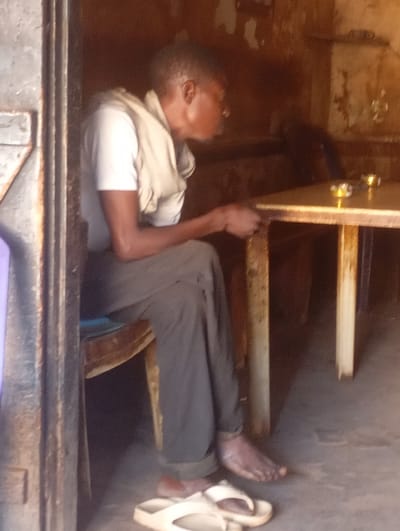 An adult male is seated at a simple wooden table in a rustic indoor setting in Yelwa, Nigeria. He is shown in profile, facing towards the right, with his hands resting near the table's edge. He wears a light-colored, short-sleeved t-shirt and dark trousers, with a light-colored fabric draped over his shoulders or neck. He is barefoot, and a pair of light-colored flip-flops are on the concrete floor near his feet.
The interior appears aged and humble, with rough, discolored walls suggesting unpainted plaster or mud construction. The wooden table has a light-colored top and dark legs. On the table, two small, shiny metallic objects, possibly cups or decorative items, are visible. The scene is illuminated by ambient daylight, creating a somewhat dim yet natural light within the room. A dark, weathered doorway or frame is prominent on the left side of the image. The man's posture suggests a state of rest or quiet contemplation. No specific activity beyond sitting is evident, though the objects on the table hint at a possible eatery or place for refreshments. No text is visible in the image.