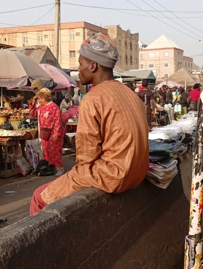 The scene depicts a bustling outdoor market in Sabon Gari West, Nigeria, under a bright daytime sky. In the immediate foreground, a dark-skinned male, facing left in profile, sits with his back mostly to the viewer on a dark, textured concrete or stone barrier. He wears a light brown, striped long-sleeved garment over dark red trousers and a grey headwrap with subtle orange patterns. To his right, a stack of white papers or thin plastic items is visible on the barrier.

In the midground, a vibrant market operates with numerous individuals and stalls. On the left, a fair-skinned woman with light hair, wearing a red patterned dress, stands amidst market goods under a light-colored umbrella. Behind her and to the right, other figures, some indistinct, populate the market area. Various goods are displayed on tables and on the ground, often under large market umbrellas of white and pink hues.

The background features several multi-story buildings, predominantly light brown or tan, under a clear sky with visible overhead utility lines. One building on the far right has a distinctive pyramidal roof structure. The overall environment indicates an active urban commercial setting.