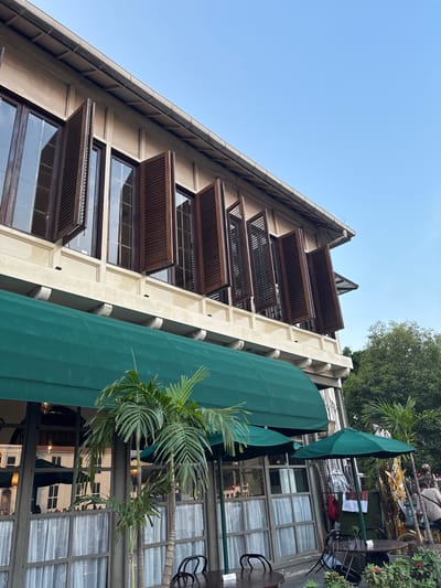The image shows the exterior of a building, likely a cafe or restaurant, in West Jakarta, Indonesia. The building features a light-colored facade with multiple large windows on the upper floor, each fitted with dark wooden shutters that are partially open. A prominent green awning extends from the building's lower level, covering outdoor seating areas. Two green umbrellas are visible, providing shade for outdoor tables. Several potted palm trees are placed in front of the building, adding to the tropical ambiance. In the background, lush green trees and a clear blue sky are visible. A few people are present in the background, suggesting a public space. The overall scene depicts a serene and inviting outdoor setting.