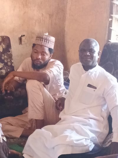 Two adult males are depicted seated indoors in Doma, Nigeria. The individual on the left has dark hair and a beard, wearing a light-colored long-sleeved tunic and a brown patterned cap. He is seated on the floor or a low cushion, with his right arm resting on his bent right knee, looking downwards. The individual on the right has a bald head and is wearing a white long-sleeved tunic with a breast pocket; he is smiling and looking towards the camera, seated on a dark cushion or chair. The background consists of a light brown wall, possibly mud-plastered, with a rectangular recess or opening behind the left individual and a window with vertical slats in the upper right. Floral patterned upholstery or fabric is visible behind the left subject. Natural light illuminates the scene, likely from the right.