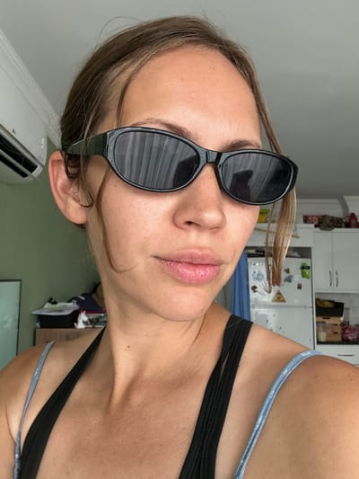 A woman is pictured from an unusual angle, wearing sunglasses. Her hair is light brown. She is wearing a black top with a blue strap. The location appears to be inside a building, potentially a residence, with a white wall and an air conditioning unit visible in the background. The scene is in Unknown, Türkiye.