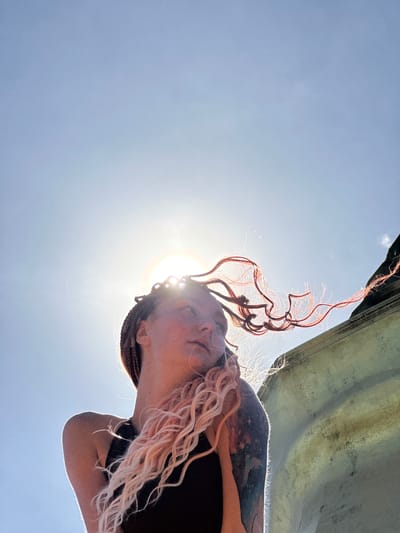 An outdoor, low-angle shot captures a woman's face and upper body against a bright blue sky. She has braids and her light pink hair is blown out in wisps by the wind, catching the sunlight. Her face is turned upward and to the side, with her eyes closed and a slight smile on her lips. She is wearing a black tank top, and a large, light-colored, weathered structure is visible to the right of the frame. The sun is prominently positioned in the upper center of the image, creating a lens flare that partially obscures her face and illuminates her hair and shoulder. The scene appears to be a sunny day, possibly on a rooftop or elevated area overlooking a city, though no specific landmarks are identifiable to confirm a location in Ko Samui, Thailand.