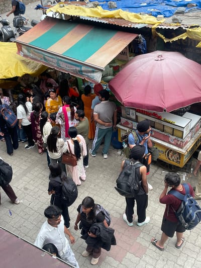 This image captures a bustling street scene in Bhagsu, India, from an elevated, downward-looking perspective. A large group of people is gathered around several informal street stalls.

In the central foreground, a street food cart with a large pink umbrella is prominent. It displays tomatoes and lemons, and features "PAYTM" QR codes for digital payments. Several individuals, including men wearing turbans, are clustered around this cart, seemingly ordering or waiting for food. Discarded plastic spoons are visible on the paved ground nearby.

To the left, another stall is covered by a striped awning in teal and muted orange/brown, with blue and yellow tarpaulins providing additional shelter. Partially visible text on a yellow banner reads "AND VEG. SH," and "AMI" is visible below the striped awning. A motorbike or scooter is parked near this area.

The crowd consists of men and women of varying ages, dressed in casual clothing. Many are carrying backpacks, suggesting they might be students, travelers, or locals engaged in daily activities. People are generally standing, some in a queue-like formation extending away from the food cart.

The setting is an outdoor, paved area, likely a busy sidewalk or market street. The lighting indicates it is daytime, possibly under an overcast sky or during the late afternoon, as shadows are soft or absent. The overall impression is one of a vibrant, active street food or market environment.